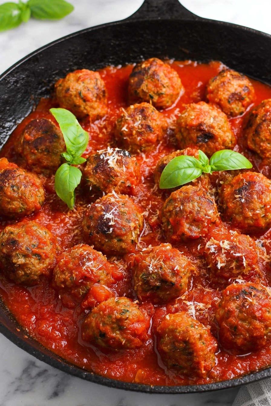 The image shows a black cast iron skillet filled with about twenty round meatballs in a thick red tomato sauce. The meatballs are textured with herbs and look cooked with slightly browned edges. The sauce covers the bottom of the skillet, soaking the meatballs partially, with some small chunks visible. There are three green basil leaves placed on top, adding a fresh contrast to the red sauce. A little grated cheese is sprinkled lightly over some meatballs. The skillet sits on a white marbled surface. Photo taken with an iphone --ar 2:3 --v 7